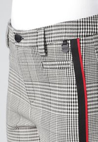 Czarno-białe spodnie w wzór pepitki z czerwono-czarnym paskiem bocznym, ozdobione czarnymi guzikami i subtelnymi detalami kieszeni.