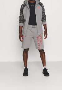 Conjunto deportivo gris que incluye una sudadera con capucha y cremallera con mangas de camuflaje, pantalones cortos a juego con texto de logo rojo y zapatillas negras. Textura suave.
