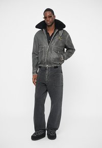 Sort denimjakke med pelskrave, lynlås lukning og forlommer, parret med løse sorte denimbukser og mønstrede fodtøj.