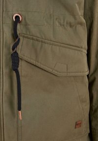 Olive green jacket featuring a textured fabric, a patch pocket with a flap, black drawstring detail, and a small brown leather logo tag.