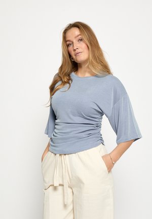 Top azzurro chiaro a maniche corte con scollo rotondo e dettagli arricciati sui lati, abbinato a pantaloni larghi color crema.