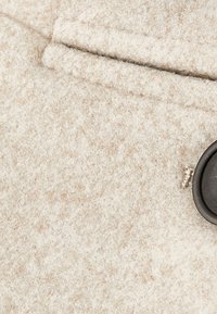 Beiger Wollstoff mit weicher Textur, versehen mit einem schwarzen Knopf und einer genähten Tasche. Dezente Farbtupfer sind im Material sichtbar.