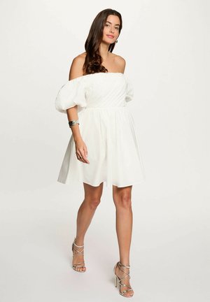 Donna che indossa un abito bianco scollato con maniche a sbuffo e sandali argento con tacco alto, in piedi davanti a uno sfondo chiaro e semplice.