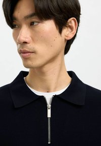 Felpa polo blu navy con mezza zip e dettaglio in metallo, tessuto a maglia texturizzato e design del colletto classico. Sotto è visibile una t-shirt bianca.