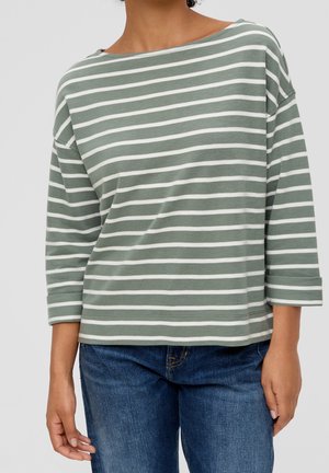 Personne portant un t-shirt à manches longues rayé horizontalement vert et blanc avec un col bateau et un jean bleu, debout devant un fond uni.