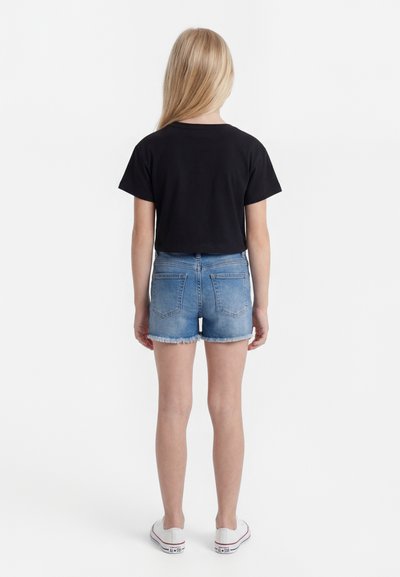 Παιδί που στέκεται με την πλάτη προς τα εμπρός, φοράει μαύρο κοντομάνικο πουκάμισο, μπλε τζιν σορτς και λευκά αθλητικά παπούτσια σε λευκό φόντο.