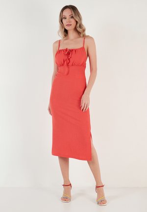 Red ribbed dress with adjustable spaghetti straps, gathered bodice, and side slit. Worn with multicolored strappy sandals.