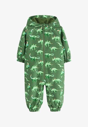 Peuter met groene dinosaurusprint jumpsuit en beige sneakers staande op een gestructureerde vloer met beige stof op de achtergrond.
