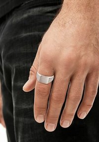 Main d'adulte masculine avec des poils clairs portant une simple chevalière en argent sur le majeur, sur un fond de pantalon foncé.