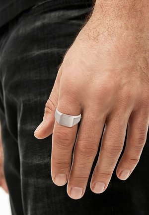 Main d'adulte masculine avec des poils clairs portant une simple chevalière en argent sur le majeur, sur un fond de pantalon foncé.