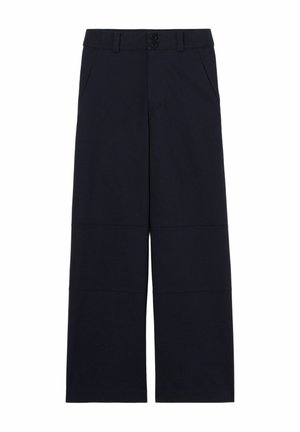Jeune femme portant un chemisier blanc, un pull jaune drapé sur les épaules, un pantalon large bleu marine, des chaussures marron et un sac à épaule blanc.