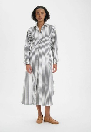 Robe chemise rayée en blanc et bleu marine, avec des manches longues, un col classique, et une longueur mi-mollet avec des fentes latérales.