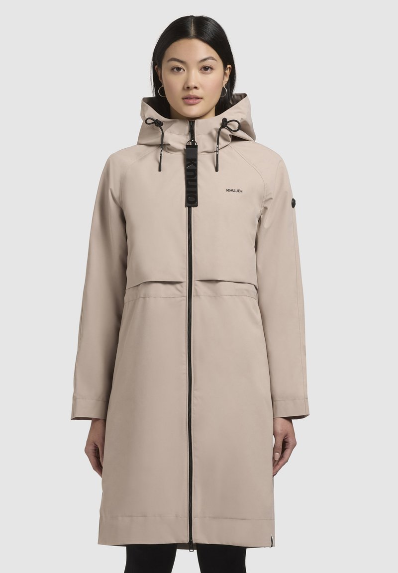 Beige waterdichte jas met een capuchon, ritssluiting aan de voorkant, elastische trekkoorden en een zwarte logoaccent op de halslijn. Glad, minimalistisch ontwerp.