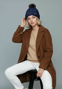 Navy knitted beanie with a pom-pom, worn with a brown coat over a beige sweater and white pants, seated on a black stool.
