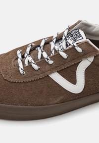 Braune Wildledersneaker mit weißem Seitestreifen, weiß-schwarz gemusterten Schnürsenkeln und sichtbarem Markenlabel auf der Zunge.