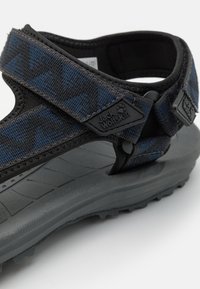 Sorte og blå mønstrete sandal med justerbare stropper, gummisåle og teksturert såle. Har merkevaren logo på stropen.