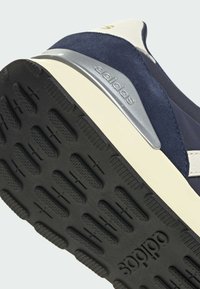 Gros plan sur le talon d'une basket Adidas bleue et blanche montrant la semelle noire texturée et le support de talon argenté avec le logo Adidas.