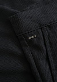 Fekete, texturált anyag közeli képe, összehajtott mandzsettával és egy kis ezüst "GABBA" márkajelzéssel a varrás közelében.