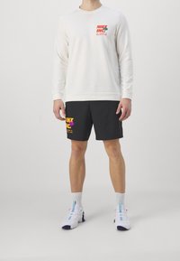 Sudadera blanca con el logo de Nike en el lado izquierdo del pecho, combinada con pantalones cortos negros que presentan un texto audaz en amarillo y rosa. Zapatillas deportivas blancas con acentos azules.