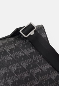 Bolsa negra con patrón de logotipo geométrico de Lacoste, material de cuero texturizado, correa ajustable y detalle de hebilla de metal plateado.