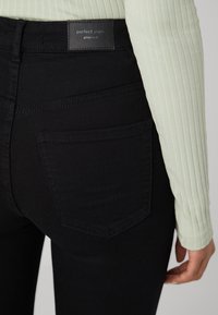 Close-up of a person wearing black high-waisted jeans labeled "perfect jeans" and a light green ribbed long-sleeve top.
