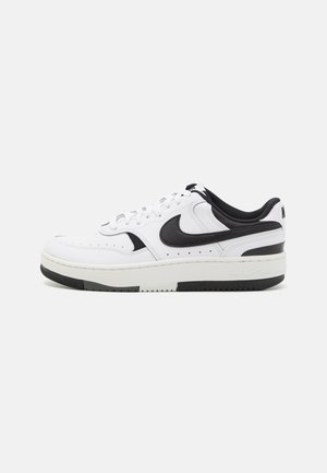 Hvid lavt snøresko med sort Nike swoosh-logo, sorte detaljer på hælen og sålen samt hvide snørebånd, vist i sideprofil på grå baggrund.