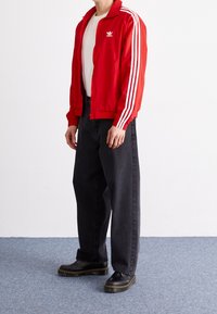 Röd jacka med vita ränder, bärs över en vit skjorta. Svarta oversized-jeans och svarta skor med gula detaljer fulländar outfiten.