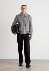 Gray zip-up jacket with two front pockets, ribbed cuffs, worn with black trousers and holding a black bag, set against a neutral background.