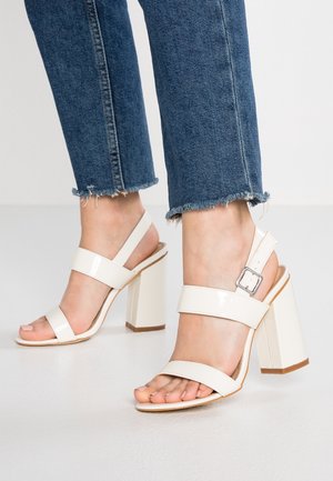 Valkoiset patenttinatuiset saappaat, joissa on leveät hihnat ja nilkkasidonta, aseteltuna repaleisten farkkujen kanssa.