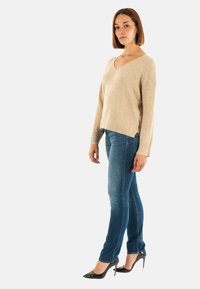 Mujer de pie de lado, vestida con un suéter beige de cuello en v, jeans ajustados azules y zapatos de tacón negro sobre un fondo blanco.