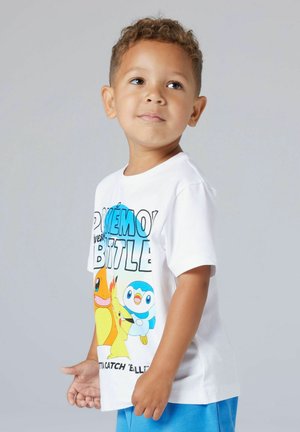 Ragazzo giovane che indossa una t-shirt bianca Pokémon Battle con Charmander, Pikachu e Piplup, in piedi davanti a uno sfondo grigio uniforme.