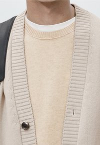 Cardigan beige en tricot avec bords côtelés, présentant un bouton sombre et un pull beige clair en dessous. Texture lisse et détail de couture subtil.