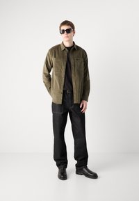 Olive green button-up shirt with two chest pockets, worn over a black t-shirt, paired with dark denim pants and black boots. Black sunglasses worn.