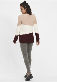 Pull en tricot avec un design de blocs de couleur en beige, blanc et bourgogne. Texture côtelée avec des manches longues, associé à un pantalon gris et des chaussures noires.