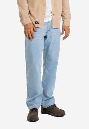 Mann trägt hellblaue Jeans, beige Hemd über weißem T-Shirt und dunkelbraune Schnürstiefel, steht vor weißem Hintergrund.