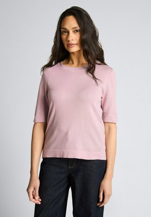 Woman with long dark hair wearing a light pink short-sleeve sweater and dark blue jeans against a plain gray background.