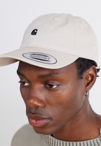 Beige justerbar caps med en sort logo. Har en buet skjerm, frontsting og en glatt bomullsoverflate.