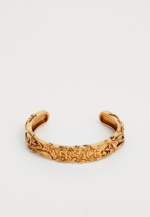 Pulsera de aro de oro con un detallado floral ornamentado, que presenta la palabra "VERSACE" en relieve en el centro. Superficie interior suave.