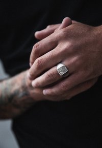 Sidabrinis žiedas su įrankiu graviruotu raštu, dėvimas ant rankos su matomais tatuiruotėmis. Paviršius turi tekstūruotą apdailą, suteikiančią tvirtą formą.