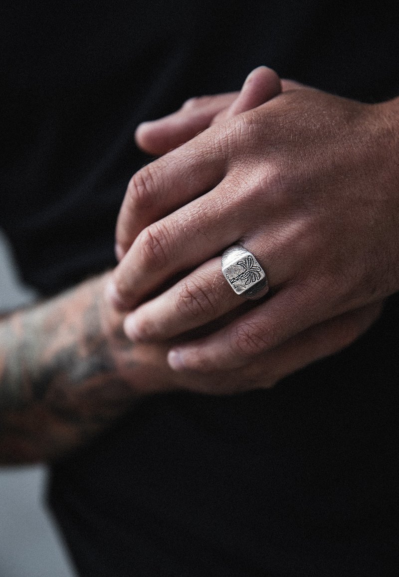 Sidabrinis žiedas su įrankiu graviruotu raštu, dėvimas ant rankos su matomais tatuiruotėmis. Paviršius turi tekstūruotą apdailą, suteikiančią tvirtą formą.