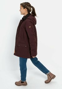 Bordeauxfarbene isolierte Jacke mit Kapuze, mit Fronttaschen und einem braunen Pelzkragen. Kombiniert mit blauen Jeans und braunen Stiefeletten.