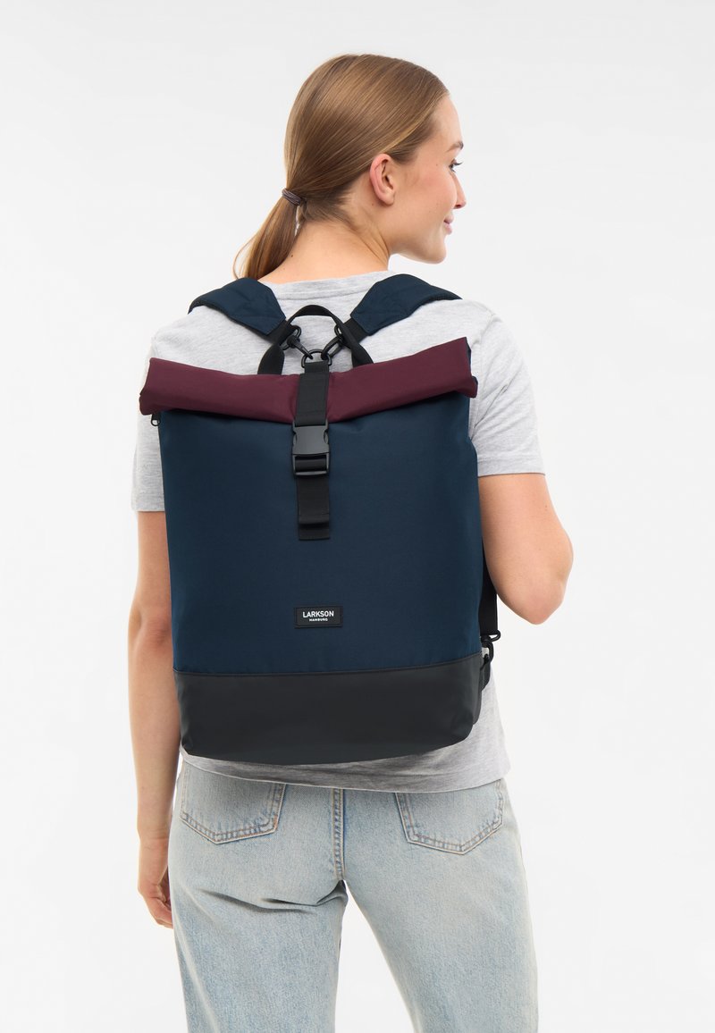 Marinblå ryggsäck med en vinröd rulltopp, svart botten, justerbara remmar och spänne. Har en logotyp på framsidan.