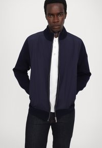 Navy bomber jacket with ribbed collar and cuffs, zip closure, featuring a smooth texture and contrasting fabric on shoulders. White polo underneath.