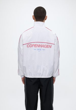 Mężczyzna w białej kurtce z czerwonymi lamówkami i napisem "COPENHAGEN" drukowanym na plecach na czerwono, w połączeniu z czarnymi spodniami z białymi paskami.