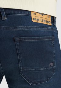 Temno modri denim kavbojki z teksturirano površino, ki vključujejo usnjeno oznako na hrbtni strani z vtisnjenim besedilom in standardno zasnovo žepov.