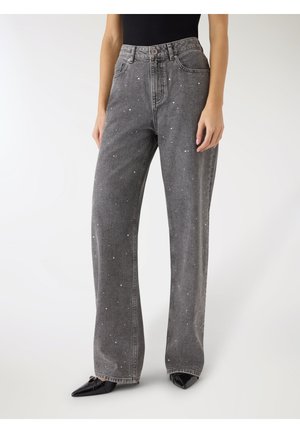 Donna che indossa jeans grigi ampi a vita alta con piccole macchie bianche e scarpe nere con punta appuntita, in piedi davanti a uno sfondo bianco.