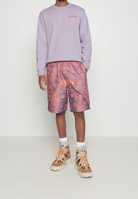 Uomo che indossa un sudore lilla, pantaloni corti a fantasia arancione e blu, calze bianche e stivali da escursionismo beige con lacci arancioni, in piedi su uno sfondo bianco.