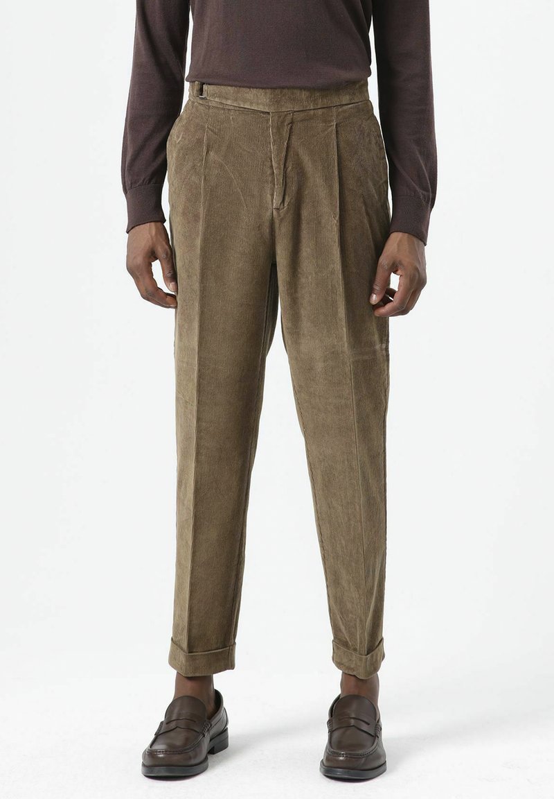 Bruine corduroy broek met geplooide voorkant, omgeslagen pijpen en zijzakken, gecombineerd met donkere schoenen en een nauwsluitende bruine top.