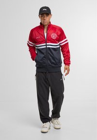 Rød og marineblå zip-up jakke med hvide striber, logo på brystet; parret med sorte bukser og hvide sneakers. Glatte, sporty materialer.