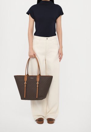 Femme portant un haut noir à manches courtes et un pantalon large crème, tenant un grand sac fourre-tout en cuir marron avec des poignées couleur tan.